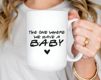 May include: White ceramic mug with the text "THE ONE WHERE WE HAVE A BABY" in black, with the word "BABY" in a colorful font. A black heart is below the text. The mug has a white handle.