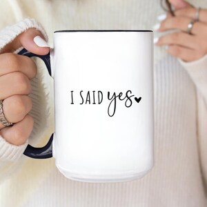 May include: White ceramic mug with a black interior and handle. The mug features the text "I SAID yes" in black script with a small heart symbol. The mug is held by a person wearing a white sweater.