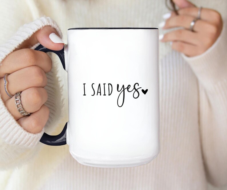 May include: White ceramic mug with a black interior and handle. The mug features the text "I SAID yes" in black script with a small heart symbol. The mug is held by a person wearing a white sweater.