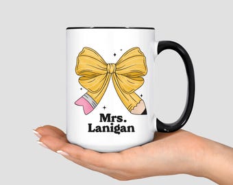 May include: White ceramic mug with a black handle and rim, featuring a yellow bow and pencil design. The mug has the text "Mrs. Lanigan" in black. The mug is held in a person's hand.
