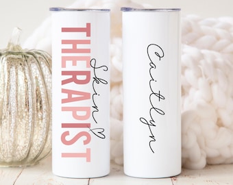 May include: Two white stainless steel tumblers. One features the text "THERAPIST Skin" in pink and black script with a heart. The other tumbler has the name "Caitlyn" in black script. Both tumblers have a silver lid.