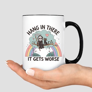 May include: White ceramic mug with a black handle and rim. The mug features a whimsical illustration of the Grim Reaper and a black cat perched on a tree branch above a rainbow. Text reads "Hang in there, it gets worse."