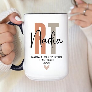 May include: White ceramic mug with black handle and rim, featuring the name "Nadia" in black script. The letters "RT" are in peach and gray, with text below reading "NADIA ALVAREZ, RT(R) RAD TECH 2025" and a small heart.