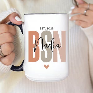 May include: White ceramic mug with black handle and rim. The mug features the text "EST. 2025 BSN Nadia" in a mix of colors, including brown, gray, and pink. A small heart graphic is below the name.