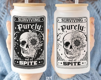 May include: Two frosted glass tumblers with bamboo lids and clear straws. Each features a black graphic of a skull adorned with sunflowers and roses, framed by decorative borders. The text reads "SURVIVING purely out of SPITE" in bold lettering.