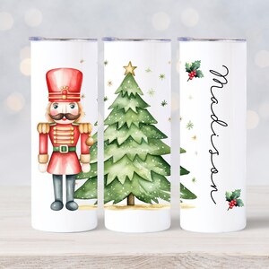 May include: Three white stainless steel tumblers with Christmas designs. The first tumbler features a red and gold nutcracker. The second tumbler features a green Christmas tree with a gold star. The third tumbler has the name "Madison" written in black cursive with a holly sprig.