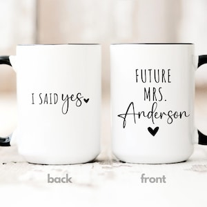 May include: Two white ceramic mugs with black handles and rims. One mug reads "I SAID yes" with a heart, the other says "FUTURE MRS. Anderson" with a heart. The mugs are for a wedding or engagement.