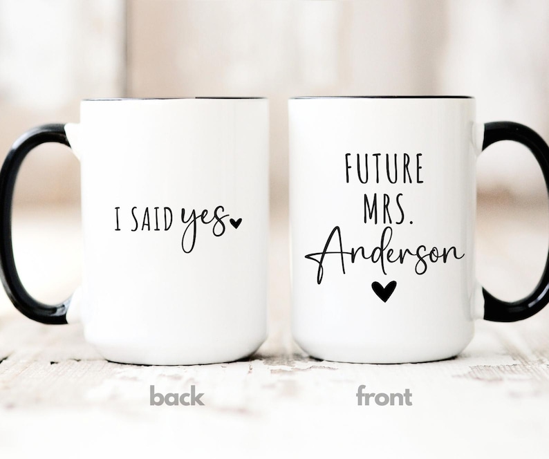 May include: Two white ceramic mugs with black handles and rims. One mug reads "I SAID yes" with a heart, the other says "FUTURE MRS. Anderson" with a heart. The mugs are for a wedding or engagement.