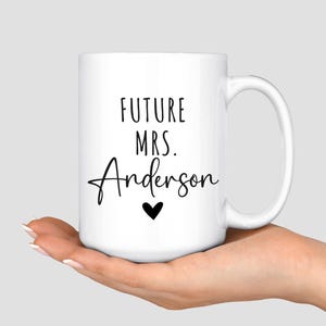 May include: White ceramic mug with the text "FUTURE MRS. Anderson" in black script, a black heart, and a white handle. The mug is held in a person's hand.