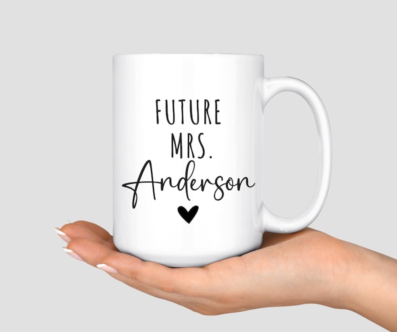 May include: White ceramic mug with the text "FUTURE MRS. Anderson" in black script, a black heart, and a white handle. The mug is held in a person's hand.