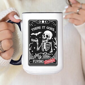 May include: White ceramic mug with a black interior and handle. The mug features a black and white graphic of a skeleton with the text "THERE IT GOES, MY LAST FLYING CENSORED". The mug is held by a person.