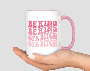 May include: White ceramic mug with a pink interior and handle. The mug features the text "BE KIND OF A BITCH" in pink, retro-style lettering. The mug is held in a hand.