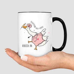 May include: White ceramic mug with a black handle and rim. The mug features a cartoon goose in a nurse's uniform holding a syringe. Text on the mug reads "Registered Silly Goose" and "Rebecca, RN".