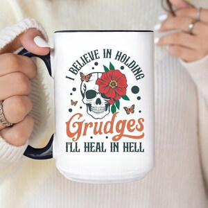May include: White ceramic mug with a black handle and rim, featuring a skull design with a red flower and butterflies. The mug has the text "I BELIEVE IN HOLDING Grudges I'LL HEAL IN HELL" in black and orange.