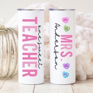 May include: Two white stainless steel tumblers with pink and black text. One tumbler says "Teacher" with the text "one sweet" written vertically next to it. The other tumbler says "Mrs. Anderson" with pink candy hearts that say "You Shine", "Ur Nice", "Awe Some", and "You Rock".