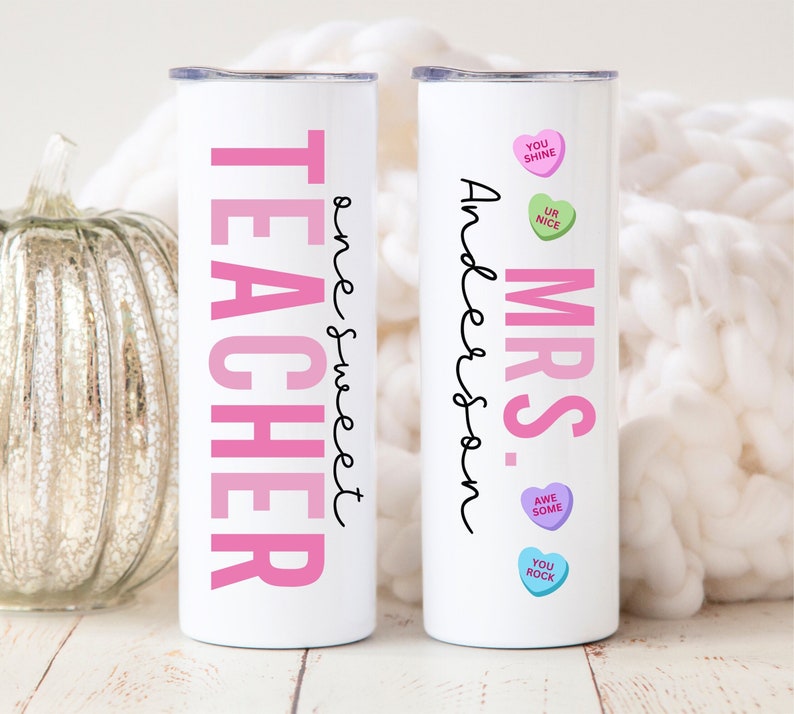 May include: Two white stainless steel tumblers with pink and black text. One tumbler says "Teacher" with the text "one sweet" written vertically next to it. The other tumbler says "Mrs. Anderson" with pink candy hearts that say "You Shine", "Ur Nice", "Awe Some", and "You Rock".