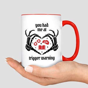 May include: White ceramic mug with a red interior and handle. The mug features a black graphic of skeleton hands forming a heart around a book, lips, and handcuffs. Text on the mug reads "you had me at trigger warning."