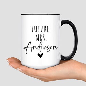 May include: White ceramic mug with a black handle and rim. The mug features the text "FUTURE MRS. Anderson" in black script, with a small black heart below the name. The mug is held in a person's hand.