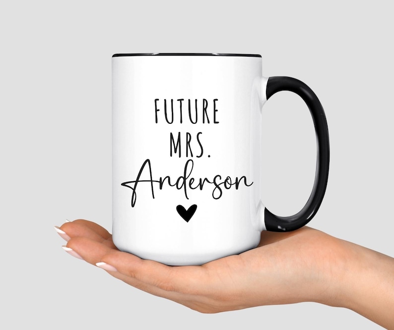 May include: White ceramic mug with a black handle and rim. The mug features the text "FUTURE MRS. Anderson" in black script, with a small black heart below the name. The mug is held in a person's hand.