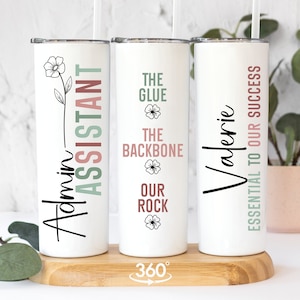 May include: Three white stainless steel tumblers with black text and floral designs. The tumblers are on a wooden base. The text on the tumblers reads "Admin Assistant", "The Glue The Backbone Our Rock", and "Valerie Essential To Our Success".