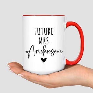 May include: White ceramic mug with a red interior and handle. The mug features the text "FUTURE MRS. Anderson" in black script, with a small black heart below the name. A simple and elegant design.