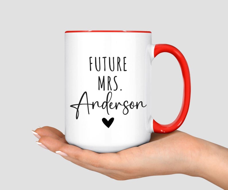 May include: White ceramic mug with a red interior and handle. The mug features the text "FUTURE MRS. Anderson" in black script, with a small black heart below the name. A simple and elegant design.