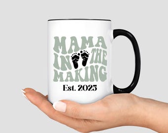 May include: White ceramic mug with a black handle and rim. The mug features the text "MAMA IN THE MAKING" in green, with black baby footprints and the year "Est. 2025".