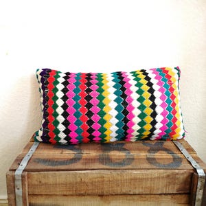 May include: A rectangular decorative pillow with a colorful geometric pattern. The pillow features a repeating diamond design in various colors, including pink, green, black, white, and yellow. It rests on a wooden crate.