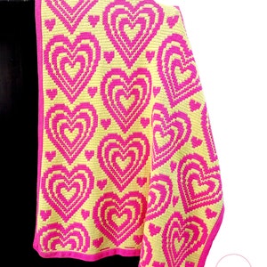 May include: A crocheted blanket with a pattern of pink hearts on a yellow background. The blanket is folded and draped over a black surface. The text "Su Patterns" is visible in the lower right corner of the image.