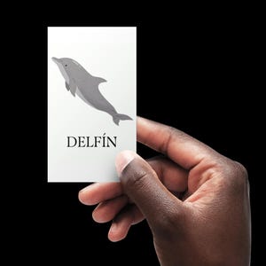 Könnte beinhalten: Eine weiße Karte mit einer grauen Delfin-Illustration und dem Wort "DELFÍN" in schwarzem Text. Die Karte wird von einer Hand vor einem schwarzen Hintergrund gehalten. Der Delfin ist stilisiert und vereinfacht dargestellt.
