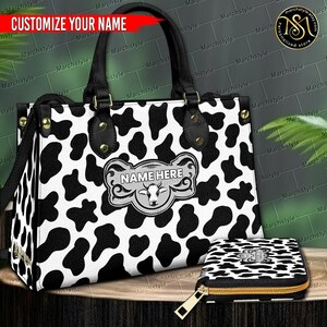 Puede incluir: Un bolso tote con estampado de vaca en blanco y negro y una cartera a juego. El bolso tote tiene asas negras y herrajes dorados. La cartera tiene una cremallera dorada. Ambos artículos tienen una placa de nombre personalizable que dice "NAME HERE".