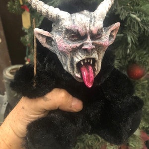 Puede incluir: Muñeco de Krampus con una cara detallada y amenazante, con cuernos, una larga lengua roja y dientes afilados. El muñeco tiene un cuerpo de piel negra y sostiene un palo de madera. El fondo incluye un árbol de Navidad con adornos.