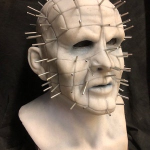 May include: A pale, sculpted mask with a grid-like pattern and numerous metal pins protruding from the head. The mask has a somber expression, with dark eye sockets and a defined mouth. The mask is a bust, resting on a white surface.