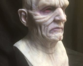 Könnte beinhalten: Eine detaillierte, realistische, weiße Silikonmaske eines Charakters mit einem faltigen Gesicht und roten Augen. Die Maske ist ein Büstenabdruck, der Kopf und Hals zeigt.