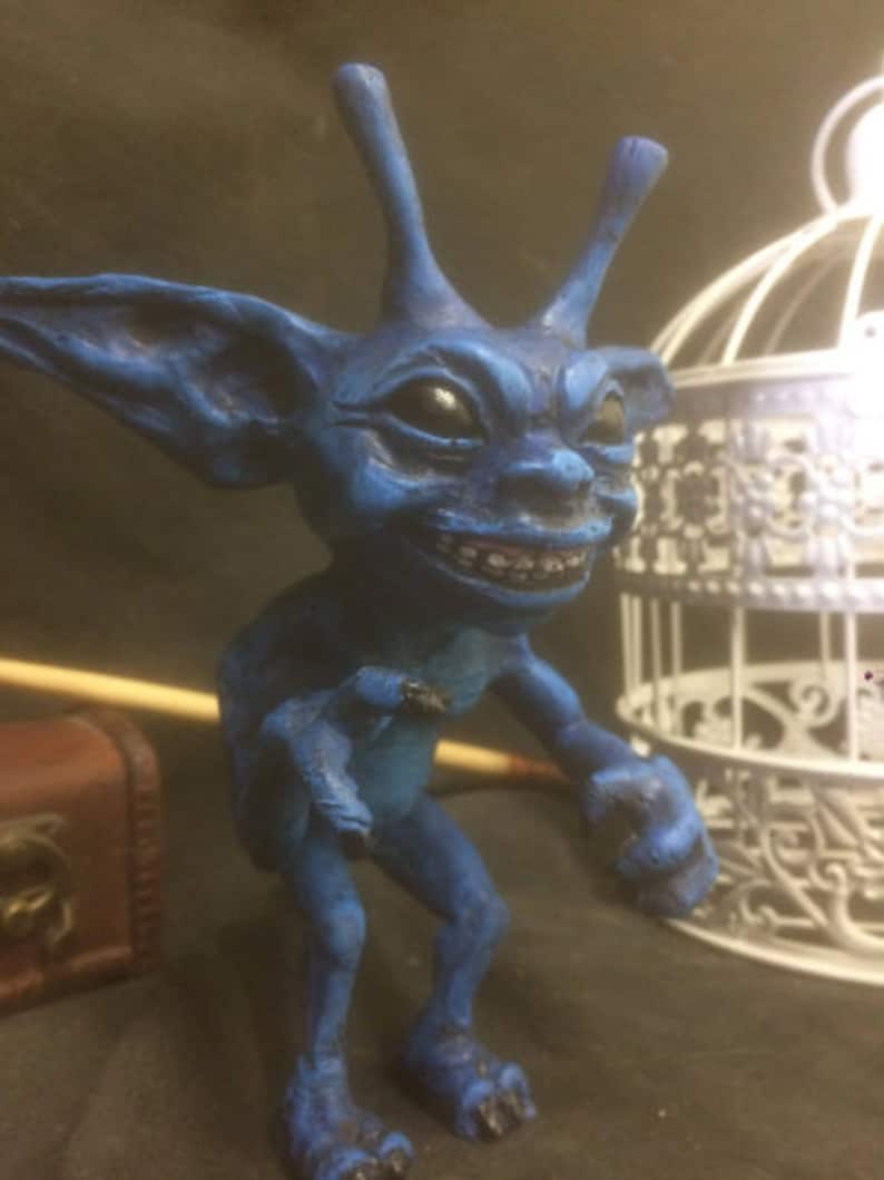 Może przedstawiać: Niebieska, ręcznie malowana figurka stworzenia przypominającego gobliny z dużymi uszami i przebiegłym uśmiechem. Figurka stoi na drewnianej powierzchni.