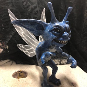 May include: A blue fairy figurine with large ears, a toothy grin, and translucent wings. The figurine is made of clay and has a whimsical, slightly menacing expression.