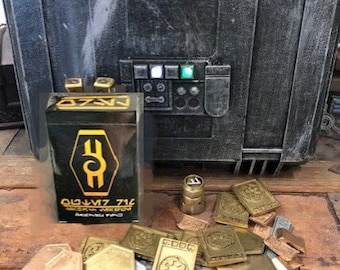 May include: A black metal case with a green and white light on the front. The case is open and a box with the text "Olm'z 716" is visible. There are also a variety of metal tokens with different designs and inscriptions.