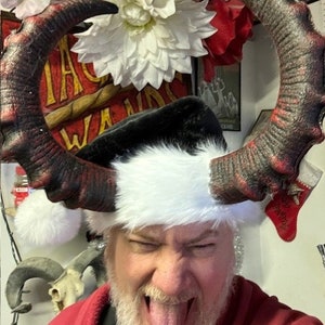 May include: A man wearing a red sweater and a Santa hat with black and red devil horns. He is sticking his tongue out and has a white beard.