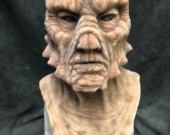 May include: A detailed, realistic, brown and tan coloured alien mask with a textured surface. The mask is made of silicone and has a human-like face with a furrowed brow, wrinkled skin, and a slightly open mouth.