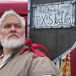 Puede incluir: Una persona con una camisa marrón con un accesorio de dragón rojo en el hombro. Detrás hay un escudo de madera con el texto "Oneail FX Studios" en letras blancas.