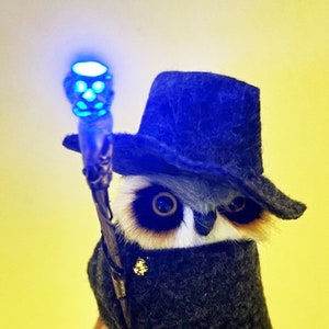 May include: A plush owl wearing a blue felt hat and holding a glowing blue and white skull-shaped staff. The owl has large brown eyes and a white face.