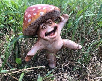 Puede incluir: Una figurita de cerámica caprichosa de un personaje de hongo con un gorro rojo y blanco, sonriendo y con los brazos extendidos. La figurita está en una posición de baile juguetona.