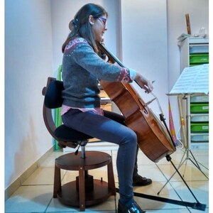 Pode incluir: Uma pessoa jovem está sentada em uma cadeira de madeira ajustável em altura com um encosto acolchoado preto e toca um violoncelo marrom. O violoncelo está apoiado em um suporte preto com um apoio para os pés.
