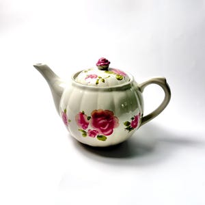 Puede incluir: Una tetera de cerámica color crema con un cuerpo acanalado en forma de calabaza y una tapa a juego. La tetera presenta un patrón floral pintado a mano de rosas rosadas con hojas verdes y un pequeño mango en forma de capullo de rosa.