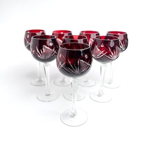 Puede incluir: Un conjunto de siete copas de vino con cuencos de color rojo intenso y patrones de cristal tallado transparente. Cada copa tiene un tallo delgado y transparente y una base circular. Los cuencos muestran grabados geométricos complejos.
