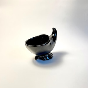 May include: A glossy black ceramic vessel with a rounded bowl shape, a single upward-curving handle, and a base featuring three distinct, claw-like feet. The object has a smooth, reflective surface and a deep, dark finish throughout its structure.