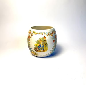 May include: A white ceramic jar with a colorful illustration of a storybook scene. The jar features a rim of fruits and foliage. The illustration depicts a child and a bird near a candle. The jar has a rounded shape.