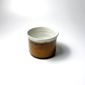 Peut inclure: Une tasse en céramique de forme cylindrique, dotée d'un extérieur émaillé brun moucheté qui se transforme en un bord foncé. L'intérieur est recouvert d'un émail blanc cassé lisse avec des stries horizontales visibles sur les parois internes.
