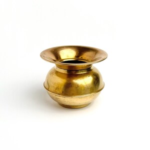 Könnte beinhalten: Ein polierter Messingtopf mit einem abgerundeten, bauchigen Körper und einem breiten, ausgestellten Rand. Die Metalloberfläche zeigt einen warmen, goldenen Farbton mit subtilen Variationen in Textur und Finish auf der Außenseite des Gefäßes.