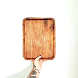 May include: A rectangular wooden serving tray with rounded corners, featuring a natural medium brown wood grain pattern. The tray has a flat base and raised edges, showcasing the organic texture and variations in the wood's color and grain.